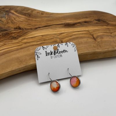 A stunning pair of Coral and Pale Red Earrings from the Ink Bloom by Claytopia collection rests gracefully on a wooden surface, accentuated by rich swirling grain patterns. These unique Inkbloom Earrings are perfect for adding a touch of elegance and style to any jewelry collection.