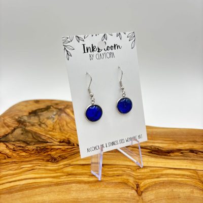 The stunning Dark Royal Blue Inkbloom Earrings are elegantly displayed on a sleek white card marked Ink Room by Clayton. Set against a polished light-grain wooden surface, these earrings deep blue hue is perfectly highlighted. This eye-catching presentation not only complements the rich color but also enhances their visual appeal. Perfect for those searching for exquisite blue earrings or luxury jewelry by Ink Room.