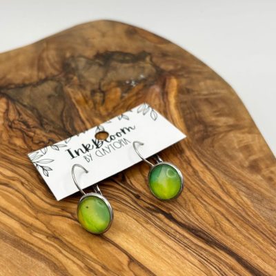 Discover the stunning Pale Green Wave Inkbloom Earrings, adorned with glossy round beads. These exquisite earrings are elegantly displayed on a wooden board, showcasing its rich swirling grain pattern, and are elegantly set against a plain white background. Perfectly hung on hooks attached to a stylish card, these earrings combine sophistication and charm in every detail.