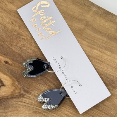 Discover stunning silver and black glittery earrings showcased on an elegant white card, adorned with the gold lettering of Spotted Petal. This exquisite display sits gracefully on a rustic wooden surface, making these dazzling earrings a must-have accessory.