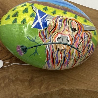 Explore a unique hand-painted oval rock showcasing a vibrant Highland cow clutching a thistle in its mouth. Set against a picturesque landscape with lush trees and the iconic blue and white Scottish flag, this eye-catching piece of art rests elegantly on a wooden surface. Perfect for those interested in Scottish-themed decor or nature-inspired art, it captures the essence of Scotlands beauty with its detailed and colorful design.