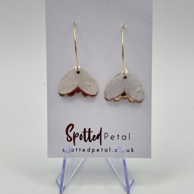 Gold hoop earrings featuring dangling white heart charms with delicate gold edges are elegantly showcased on a Spotted Petal branded card. The display stands on a transparent holder, set against a simple backdrop.
