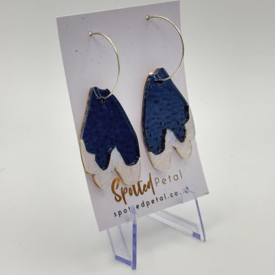 A stunning pair of earrings featuring blue and white petal-shaped pendants is elegantly showcased on a display card labeled Spotted Petal. These fashionable earrings with circular hooks are presented on a clear stand, set against a simple background, making them the perfect accessory for any jewelry aficionado. Ideal for those searching for trendy, petal-inspired earrings, these eye-catching pieces effortlessly blend style and elegance.