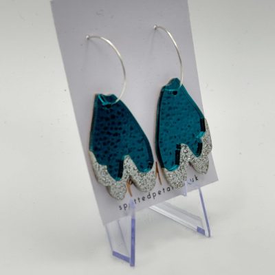 Discover a stunning pair of teal and silver glitter earrings, beautifully crafted in the shape of stylized leaves. These dazzling leaf-patterned earrings feature a textured design and are elegantly displayed on a clear stand. Perfect for adding a touch of sparkle to any outfit, they come attached to a chic card showcasing the brand name spiritedpetals.uk. Enhance your jewelry collection with these unique and eye-catching accessories today!.