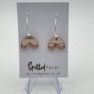 Heart-shaped pendant earrings with a blend of rose gold and glittering accents are beautifully displayed on a white Spotted Petal card. These eye-catching earrings, available at spottedpetal.co.uk, are elegantly showcased on a clear stand for perfect presentation.