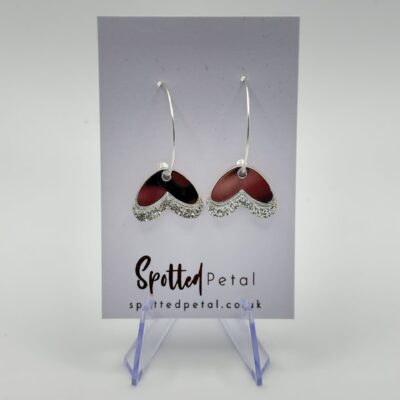 Heart-Shaped Silver Hoop Earrings with Red Enamel Center Displayed on Spotted Petal Card. Discover these elegant silver heart hoop earrings featuring a vibrant red enamel center, beautifully showcased on the branded Spotted Petal card. The clear stand and plain white background enhance their stunning design, making them the perfect accessory for any jewelry lover.