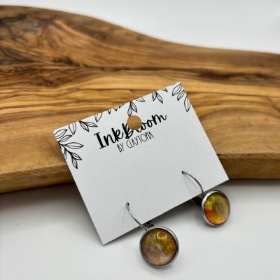 Capture the elegance of these Metallic Inkbloom Earrings, featuring vibrant red accents and an abstract design. Displayed on a stylish Inkbloom by Clayform card, this close-up showcases the intricate craftsmanship against a rustic wooden surface with natural grain. Perfect for those seeking unique, handmade jewelry pieces to elevate their style.