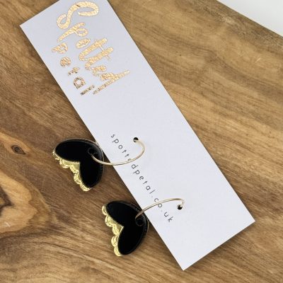Black heart-shaped earrings featuring elegant gold accents are beautifully displayed on a white card imprinted with the Spotty Pear logo in luxurious gold foil. This chic presentation rests on a rustic wooden surface, adding a touch of natural charm. Perfect for your trending jewelry collection!.