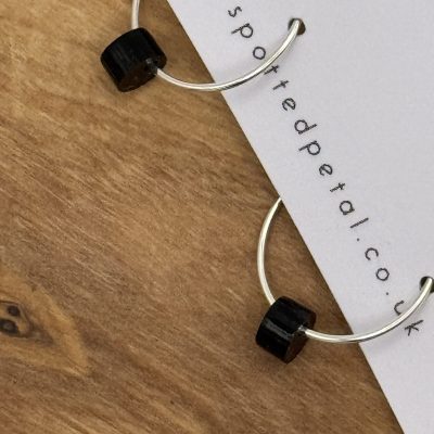Enhance your jewelry collection with stunning silver hoop earrings featuring sleek black cylindrical beads from SpottedPetal.co.uk. These elegant earrings are presented on a chic white card, beautifully displayed against a rustic wooden background. Perfect for adding a touch of sophistication to any outfit, these handcrafted earrings are a must-have accessory.