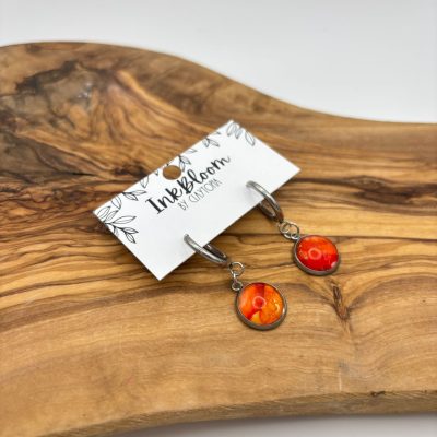 The Deep Orange Wave Hoop Inkbloom Earrings, crafted with silver hoops and eye-catching orange and red pendants, are beautifully displayed on a wooden surface. These artistic earrings are attached to an InkBloom by Kamlyn card, showcasing their modern elegance and unique style. Perfect for those seeking vibrant jewelry that combines creativity with contemporary fashion.