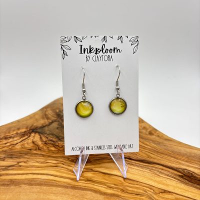Featured on the card Inkbloom by Claytopia, the Pale Yellow Green Inkbloom Earrings showcase round stainless steel accents that beautifully contrast against a wooden surface and white background. Perfect for those seeking unique, eco-friendly jewelry design.