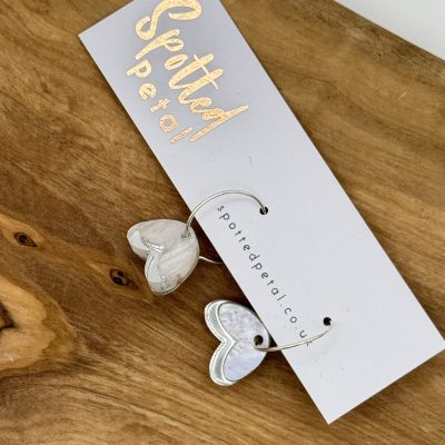 A stunning pair of heart-shaped earrings with a lustrous pearly finish is displayed on a white card labeled Spotted Petal. These exquisite earrings offer an elegant touch to any ensemble. Photographed on a rustic wooden surface, this jewelry piece stands out as the perfect gift for fashion enthusiasts or anyone looking to enhance their collection with unique and stylish accessories.
