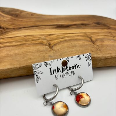 Introducing the stunning Pale Green Wave Inkbloom Earrings, designed to captivate with vibrant orange and red circular pendants. These earrings showcase an elegant wave pattern and are perfect for adding a touch of color to any outfit. Displayed beautifully on a white presentation card featuring a floral border, labeled InkBloom by Claytopia, these earrings are set against an inviting wooden surface that enhances their natural beauty. Elevate your style effortlessly with these eye-catching earrings today!.