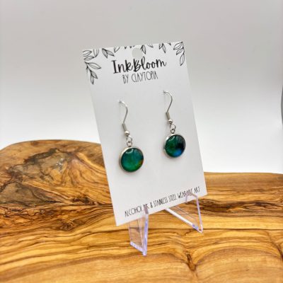 Discover the stunning Sea-green Inkbloom Earrings, featuring a captivating blue round design and delicate hooks. Elegantly showcased on a pristine white Inkbloom by Claytopia card, these exquisite earrings are beautifully presented against a light wood background. Perfect for those seeking stylish and unique handmade jewelry.