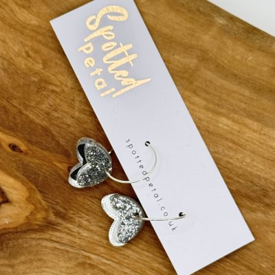 Discover the elegance of silver glitter heart-shaped earrings showcased on a white Spotted Petal card. These stunning earrings, perfect for adding sparkle to any outfit, are beautifully displayed on a wooden surface. Find more stylish jewelry pieces by visiting the URL provided on the card.