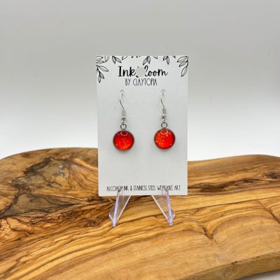 Discover our stunning Speckled Orange Inkbloom Earrings with a lustrous finish, elegantly dangling from silver hooks. These exquisite earrings are beautifully showcased on an Ink Room by Clayton card, perched on a sleek clear stand over a uniquely grained wooden surface. Perfect for adding a touch of elegance to any outfit!.