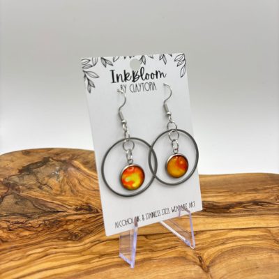 The Statement Orange and Yellow Circle Earrings from Inkbloom by Claynora are beautifully displayed on a branded card. These bold earrings feature vibrant pendants hanging from silver hoops, elegantly supported by a transparent stand on a wooden surface.