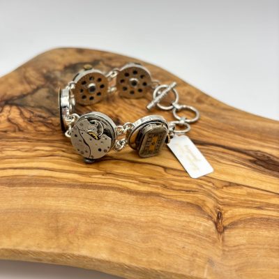 A stunning silver bracelet crafted from vintage watch movements elegantly rests on a wooden surface. This unique piece features an array of round and rectangular watch components meticulously connected by silver rings, highlighting intricate gears and detailed craftsmanship.