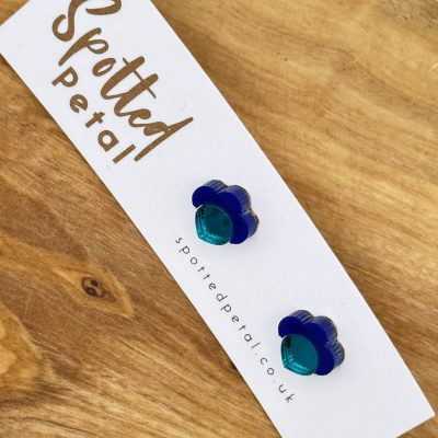 Discover the elegance of blue and teal flower-shaped earrings from Spotted Petal. These stunning earrings, elegantly displayed on a branded card, rest beautifully on a wooden surface. Perfect for adding a touch of floral charm to your accessory collection.