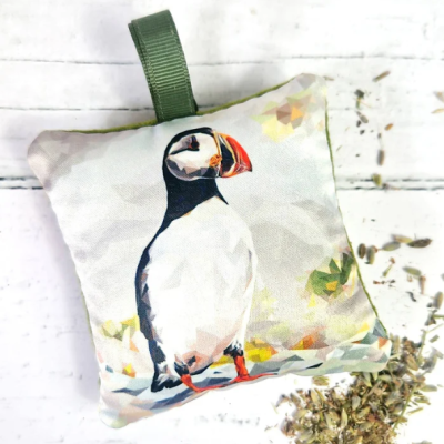 Experience the charm of The Lavender Sachet - Atlantic Puffin, featuring a decorative pillow showcasing an artistic representation of the Atlantic Puffin. This unique design utilizes geometric shapes and muted colors for a modern touch. Complemented by a green hanging loop, set against a crisp white background with dried herbs, this sachet creates a rustic lavender ambiance perfect for invigorating any space with natural freshness. Ideal for those seeking nature-inspired home decor with an eye-catching Atlantic Puffin image.