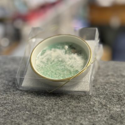 A decorative small ceramic bowl with an elegant gold rim, containing light green healing crystals and soft white feather-like filler, rests on a gray surface inside a transparent plastic display box. The background is artfully blurred to highlight the bowls unique details—perfect for home decor, crystal collectors, or as a thoughtful gift item.