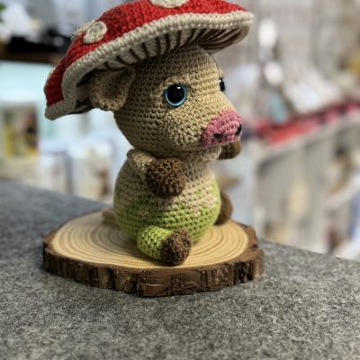 Handcrafted Crocheted Plush Toy with Cow Body and Red Mushroom Cap with White Spots, Featuring Big Blue Eyes. Perfectly Displayed on a Wooden Disc in Craft Store on Gray Surface.