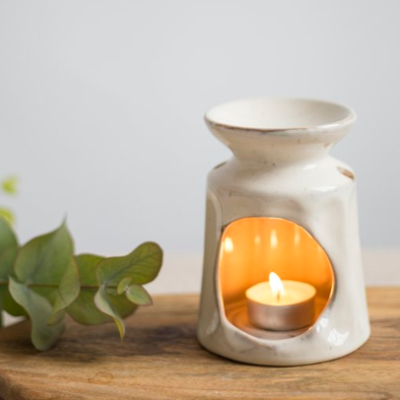 A glowing tealight candle illuminates a beige wax melt burner with an open-front design, elegantly positioned on wooden surfaces. Complemented by fresh eucalyptus leaves, this setup creates a peaceful ambiance against a softly blurred gray background.