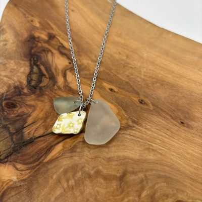 Silver chain necklace featuring two frosted sea glass pendants and a unique yellow floral ceramic shard pendant, elegantly displayed on a smooth natural wooden board. Perfect for ocean-inspired jewelry lovers seeking handmade artisan necklaces with coastal charm.