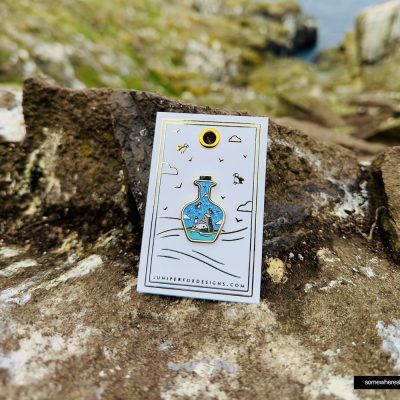 The Puffin in a Bottle enamel pin showcases a captivating scenic landscape encapsulated within a bottle, perfectly set against rocky terrain. Accompanied by a Juniper Fox Designs card, this package enhances the sense of mystery with its blurred depiction of rocks and water. Ideal for nature enthusiasts and collectors alike, this unique puffin enamel pin is both an enchanting accessory and a charming gift option.
