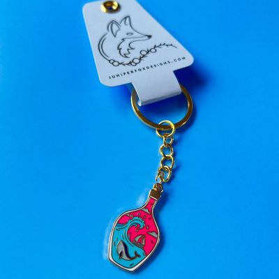 Enhance your accessory collection with a vibrant keychain featuring a potion bottle design in abstract pink and teal colors. This unique keychain hangs from a stylish gold ring, complete with a tag featuring a distinctive wolf logo. Visit juniperfoxdesigns.com for more creative designs, showcased against an eye-catching bright blue background.
